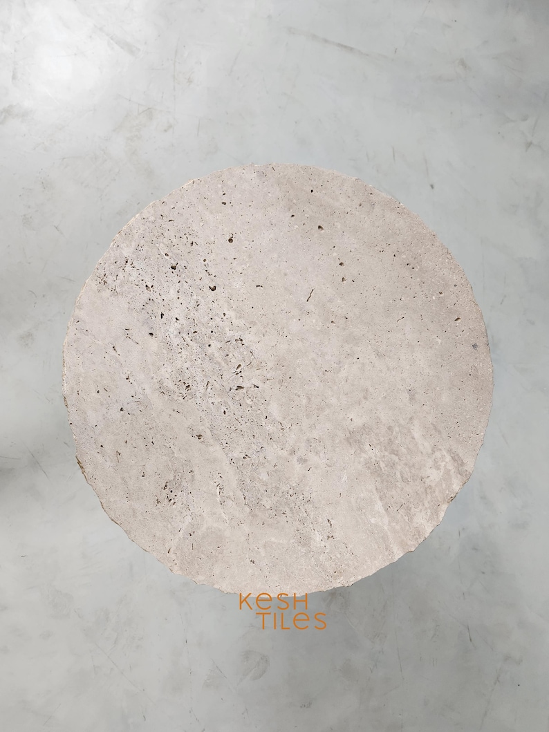 May include: A round, light beige travertine stone table top with a rough, textured surface. The table top is approximately 18 inches in diameter.