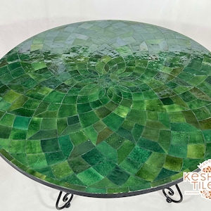 Puede incluir: Mesa redonda de mosaico con un patrón verde y verde oscuro. La mesa tiene una base de metal negro con adornos decorativos. La parte superior del mosaico está hecha de pequeñas baldosas cuadradas dispuestas en un diseño radial.