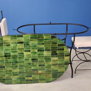 Puede incluir: Una mesa ovalada de mosaico verde con marco de metal negro, acompañada de dos sillas a juego con cojines blancos. La superficie de la mesa es un mosaico de azulejos verdes. Las sillas tienen un diseño curvo.