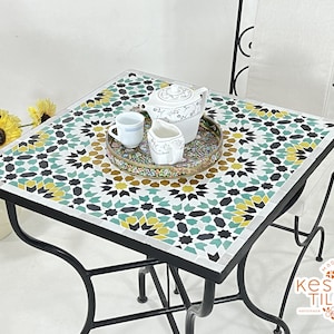 May include: A black metal table with a square top made of colorful mosaic tiles. The table has a geometric pattern in shades of green, black, white, and yellow. A white teapot, creamer, and cup are on the table.