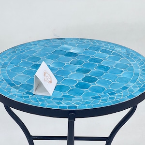Puede incluir: Una mesa redonda de mosaico azul con un marco de metal negro. La superficie de la mesa está hecha de pequeñas baldosas azules irregulares.