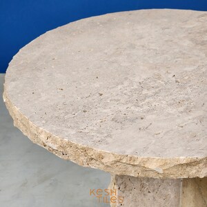 May include: A round, light beige travertine table with a rough, uneven edge. The table has a thick, sturdy base and is made of natural stone.
