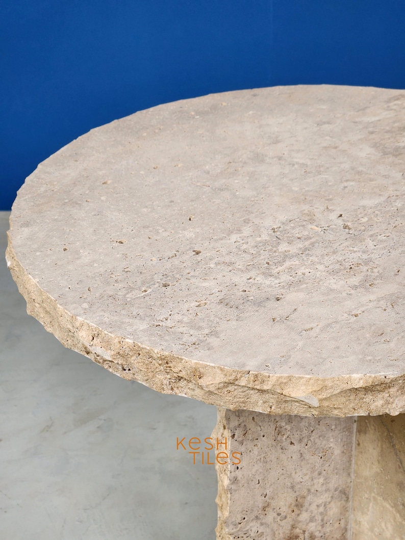 May include: A round, light beige travertine table with a rough, uneven edge. The table has a thick, sturdy base and is made of natural stone.