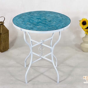 May include: A round mosaic tile table with a white metal frame. The tabletop features a teal blue mosaic tile design. A decorative gold lantern and a vase of sunflowers are on either side of the table.