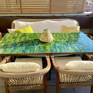 Peut inclure: Ensemble de salle à manger d'extérieur avec une table rectangulaire à mosaïque verte et bleue. Deux chaises avec dossiers en osier et coussins crème sont placées devant la table. Une lampe tressée et un canapé blanc avec des coussins jaunes complètent la scène. La table mesure environ 150 cm de long.
