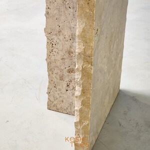 May include: Close-up of the corner of a light beige travertine stone table. The table has a rough, textured surface and a slightly irregular shape.