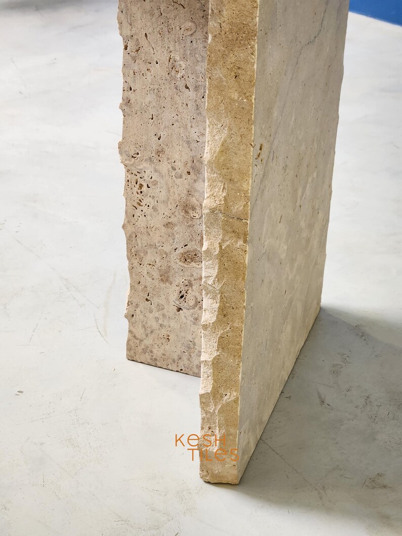 May include: Close-up of the corner of a light beige travertine stone table. The table has a rough, textured surface and a slightly irregular shape.
