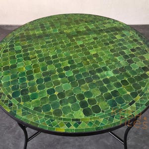 May include: A round table featuring a mosaic top composed of numerous small, square and octagonal green tiles in varying shades. The tabletop is framed by a decorative border of triangular and diamond-shaped tiles, supported by a black metal frame.