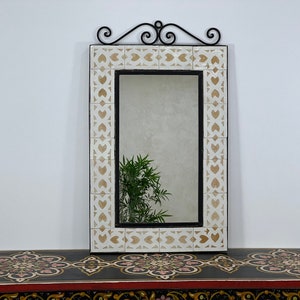 Peut inclure: Un miroir en céramique blanche avec un cadre en métal noir et un motif de volutes décoratif en haut. Le miroir a un motif géométrique autour de la bordure.