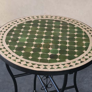 May include: A round table featuring a mosaic top with green octagonal tiles and a border of cream-colored triangular and rectangular tiles. The table is supported by a black metal frame with curved legs and a decorative scrollwork base.