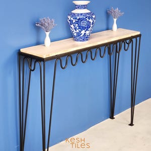 Puede incluir: Una mesa consola de mármol blanco con patas de horquilla negras. La mesa tiene un diseño ondulado en el borde inferior de la parte superior. Un jarrón de porcelana azul y blanco con diseños florales se encuentra sobre la mesa.