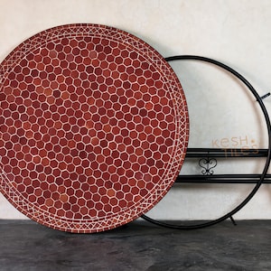 May include: A round table top featuring a red hexagonal tile mosaic pattern with a white grout border and a decorative lattice edge. Beside it stands a black metal table base with a circular frame and a small heart-shaped metal detail.