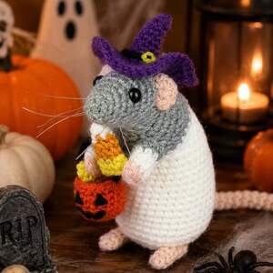 Könnte beinhalten: Eine gehäkelte graue und weiße Rattenfigur mit einem violetten Hexenhut mit gelbem Band. Die Ratte hat schwarze Augen, rosa Ohren und Füße und hält einen kleinen orangefarbenen Kürbiseimer, der mit gelben, orangen und weißen Zuckermaisstücken gefüllt ist.