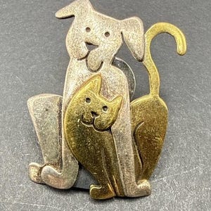 Peut inclure: Une broche en métal représentant un chien argenté assis avec un chat doré placé devant lui. Le chien a des oreilles tombantes et une expression faciale simple, tandis que le chat a des oreilles pointues et une queue courbée vers le haut.