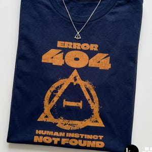 Puede incluir: Una camiseta azul marino con el texto "ERROR 404 HUMAN INSTINCT NOT FOUND" en naranja. Un collar con colgante de triángulo plateado está sobre la camiseta. La camiseta tiene un diseño gráfico de un triángulo con la letra "I" en su interior.