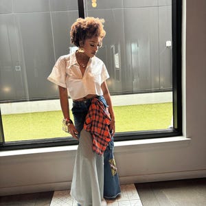 May include: A woman wearing a white cropped button-down shirt with a plaid shirt tied around her waist. She is wearing blue jeans with a patchwork design and a light blue skirt layered over the jeans.
