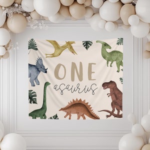 May include: A beige backdrop with the text "ONEsaurus" in brown lettering. The backdrop is decorated with green leaves and various colorful dinosaurs, including a green dinosaur, a blue dinosaur, a brown dinosaur, and a yellow dinosaur.