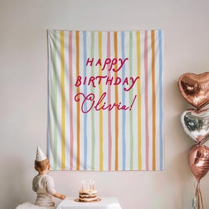 May include: A birthday backdrop with pastel-coloured vertical stripes. The text "Happy Birthday Olivia!" is written in pink. Three heart-shaped balloons and a small cake with candles are also visible. The backdrop is a decorative item.