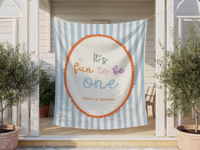 May include: A banner with a light blue and white striped background. A white circle with an orange scalloped border in the center features the text "It's fun to be one" in colorful letters. The text "Ezra's 1st Birthday" is below.