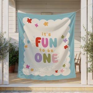 May include: A light blue banner with the text "it's FUN to be ONE" in colorful letters. The banner has a white scalloped border with colorful flower accents. The banner is hanging outside.