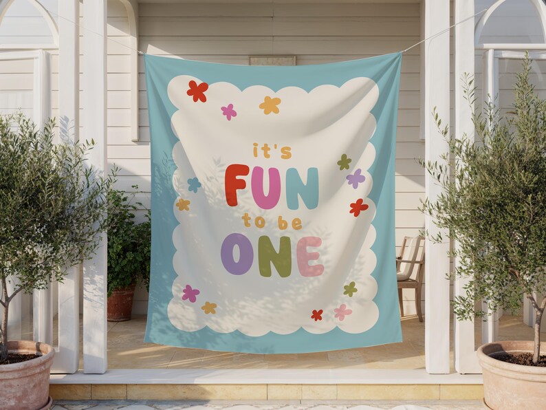 May include: A light blue banner with the text "it's FUN to be ONE" in colorful letters. The banner has a white scalloped border with colorful flower accents. The banner is hanging outside.