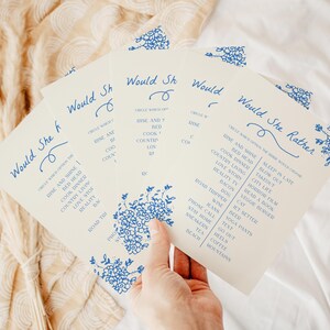 May include: A set of off-white paper game cards titled Would She Rather in blue ink. The cards feature a floral border and a list of choices including Rise and Shine, Bed Head, Cook Dinner, Country Living, Love Story, Reality TV, and Bacon.