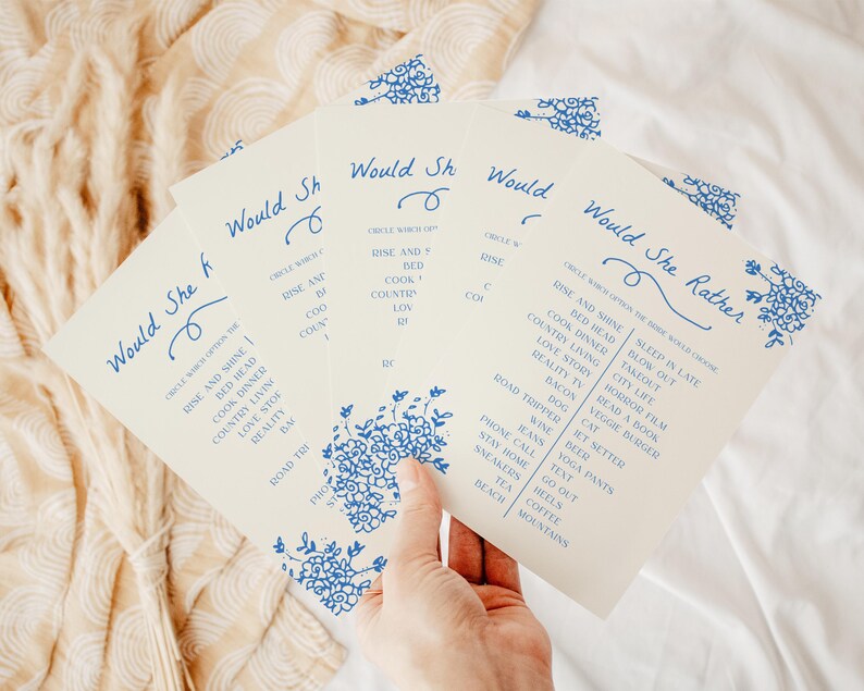 May include: A set of off-white paper game cards titled Would She Rather in blue ink. The cards feature a floral border and a list of choices including Rise and Shine, Bed Head, Cook Dinner, Country Living, Love Story, Reality TV, and Bacon.