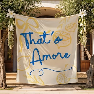 May include: A cream banner displaying the words "That's Amore" in blue script, with yellow lemon and floral designs. The banner is hung between two trees, decorated with white flowers and greenery, creating a celebratory outdoor scene.