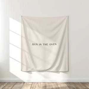 May include: Cream-colored wall tapestry with the phrase "BUN IN THE OVEN" printed in a dark brown font. The tapestry is hanging on a white wall, with a wooden floor visible below. The fabric has soft folds.