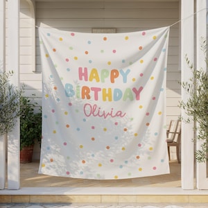 May include: A white banner with colorful polka dots and the words "HAPPY BIRTHDAY Olivia" in pastel colors. The banner is hanging outside, with plants and a chair visible in the background. A festive decoration for a birthday celebration.