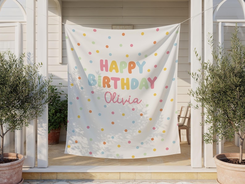 May include: A white banner with colorful polka dots and the words "HAPPY BIRTHDAY Olivia" in pastel colors. The banner is hanging outside, with plants and a chair visible in the background. A festive decoration for a birthday celebration.