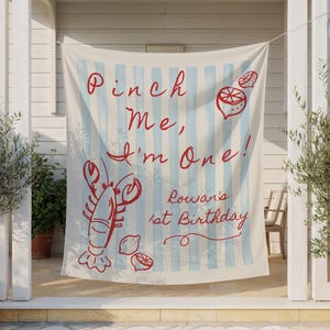 Op de afbeelding: Een cr&egrave;mekleurige banner met lichtblauwe verticale strepen en rode tekst met de tekst "Pinch Me, I'm One!" met illustraties van een kreeft en citroen. De banner zegt ook "Rowan's 1st Birthday".