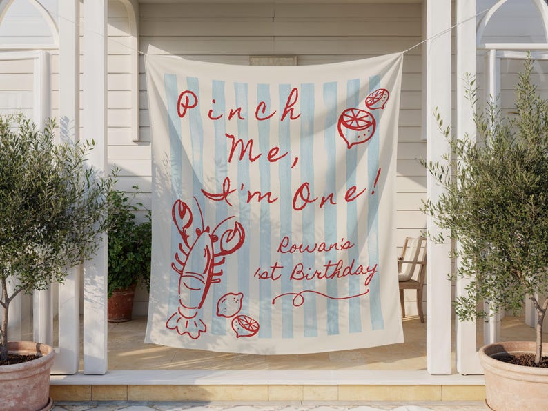 Op de afbeelding: Een cr&egrave;mekleurige banner met lichtblauwe verticale strepen en rode tekst met de tekst "Pinch Me, I'm One!" met illustraties van een kreeft en citroen. De banner zegt ook "Rowan's 1st Birthday".