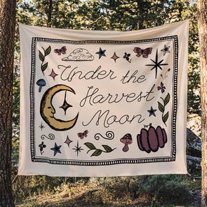 May include: A square off-white fabric wall hanging featuring the text "Under the Harvest Moon" in black script. The design includes a yellow crescent moon with a face, a purple pumpkin, butterflies, stars, mushrooms, and green foliage.