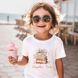 May include: A white t-shirt featuring a watercolour illustration of a stack of books with floral details and the text "Chapter Two". The person in the image is wearing sunglasses and holding a pink ice cream cone.