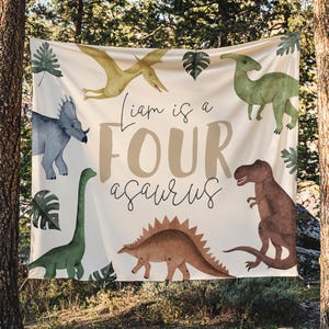 May include: A beige banner with dinosaur illustrations and the text "Liam is a FOUR asaurus". The banner features a variety of dinosaurs in shades of green, brown, and blue, with green leaf accents.