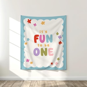 May include: A wall tapestry with a light blue border and a white center. The text "it's FUN to be ONE" is displayed in colorful letters, surrounded by small flower illustrations. The tapestry is hanging on a white wall.