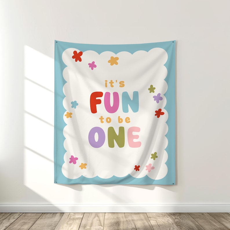 May include: A wall tapestry with a light blue border and a white center. The text "it's FUN to be ONE" is displayed in colorful letters, surrounded by small flower illustrations. The tapestry is hanging on a white wall.