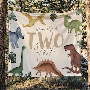 May include: A beige banner with dinosaur illustrations and the text "Liam is a TWO Rex." The banner features a variety of dinosaurs in shades of green, brown, and blue, along with tropical leaves.