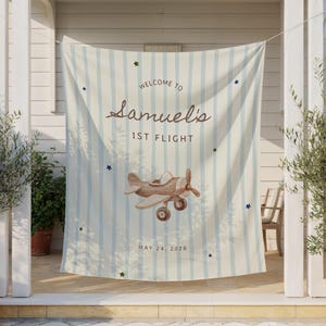 May include: A light blue and white striped banner with the text "Welcome to Samuel's 1st Flight" and a brown airplane illustration. The banner also includes the date "May 24, 2028" and star accents. The banner is hanging outside.