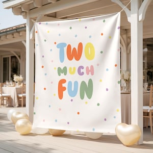 May include: A white banner with the words "TWO MUCH FUN" in colorful bubble letters and scattered polka dots. The banner is hanging outdoors, with gold balloons and a heart-shaped balloon at the bottom, suggesting a birthday party theme.
