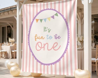 May include: A banner with pink and white vertical stripes, featuring an oval with the text "It's fun to be ONE" in colourful lettering. The oval is outlined in purple with a scalloped edge and topped with a string of colourful pennants. Gold balloons are in the foreground.