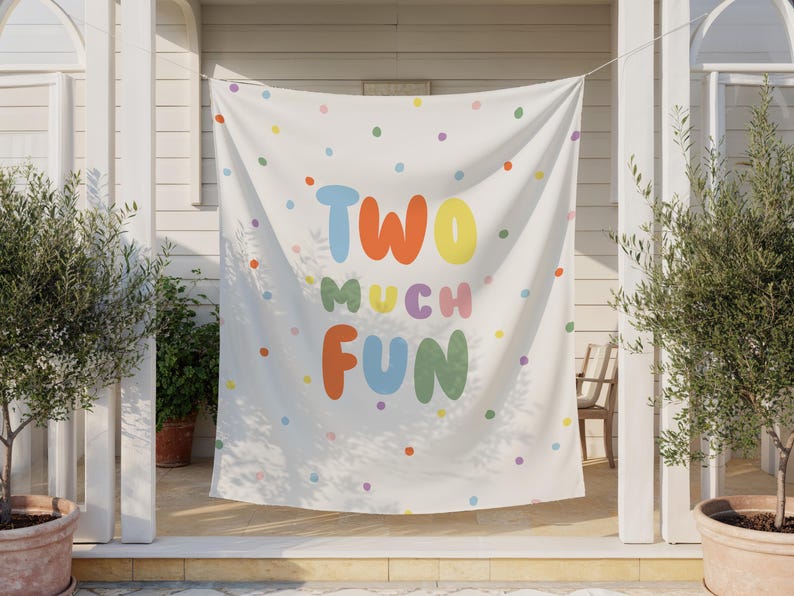 May include: A white banner with colorful polka dots and the words "TWO MUCH FUN" in large, playful letters. The text is in various colors, including blue, orange, yellow, purple, and green. The banner is hanging outdoors.