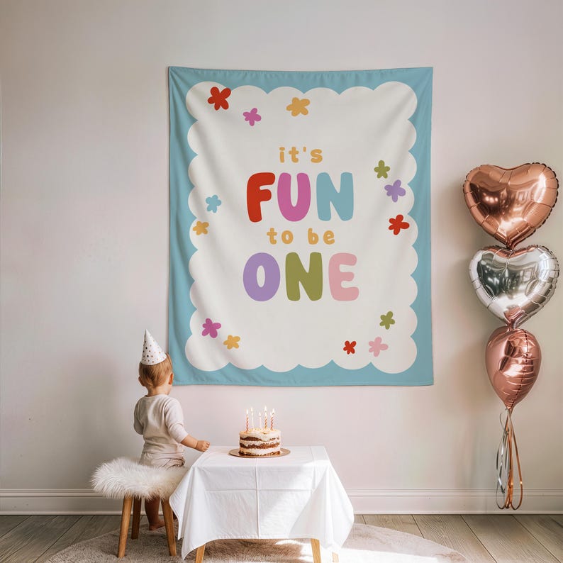 May include: A birthday party scene with a large banner that reads "it's FUN to be ONE." The banner is decorated with colorful flowers and a scalloped edge. A small cake with lit candles sits on a table, and heart-shaped balloons are visible.