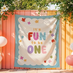May include: A colorful banner with the text "it's FUN to be ONE" in bold letters, surrounded by a scalloped border and floral accents. Balloons in various colors decorate the scene, creating a festive atmosphere.