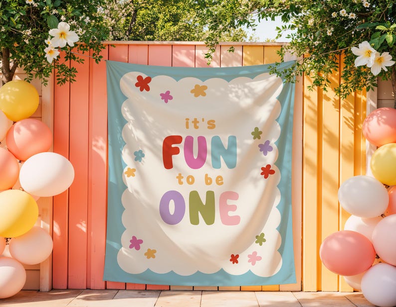 May include: A colorful banner with the text "it's FUN to be ONE" in bold letters, surrounded by a scalloped border and floral accents. Balloons in various colors decorate the scene, creating a festive atmosphere.