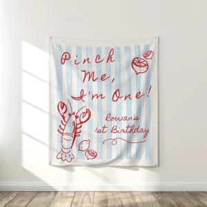 Op de afbeelding: Een wandkleed met een lichtblauwe en witte gestreepte achtergrond. Rode illustraties van een kreeft, citroenen en de tekst "Pinch Me, I'm One! Rowan's 1st Birthday" staan op de stof.