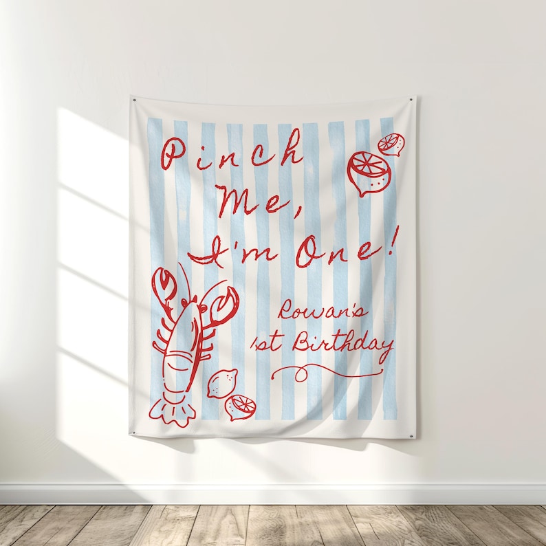 Op de afbeelding: Een wandkleed met een lichtblauwe en witte gestreepte achtergrond. Rode illustraties van een kreeft, citroenen en de tekst "Pinch Me, I'm One! Rowan's 1st Birthday" staan op de stof.