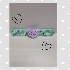 May include: A handmade crochet headband in a light teal color, with a lavender heart in the center. The headband is a soft, textured design, perfect for adding a touch of handmade charm to any outfit. The heart adds a sweet, romantic touch.
