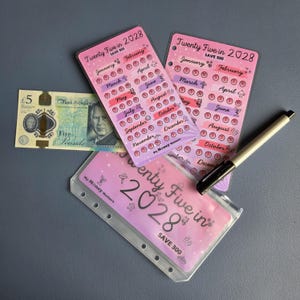 May include: Two pink plastic savings tracker inserts and one clear binder pouch labelled Twenty Five in 2028 SAVE 300, featuring monthly circles for tracking money, alongside a five pound note and a black and gold pen for financial planning.